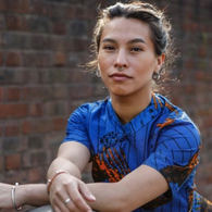 A young woman with dark hair tied back, wearing a blue patterned shirt, sitting outdoors against a brick wall.