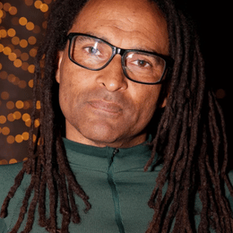 A man with long dreadlocks wearing glasses and a dark green shirt, posing against a dark background with orange lights.