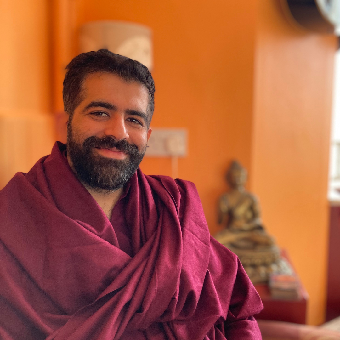 A man with a beard and short dark hair smiling, wearing a maroon robe, sitting indoors with an orange wall, a Buddha statue, and a window in the background.