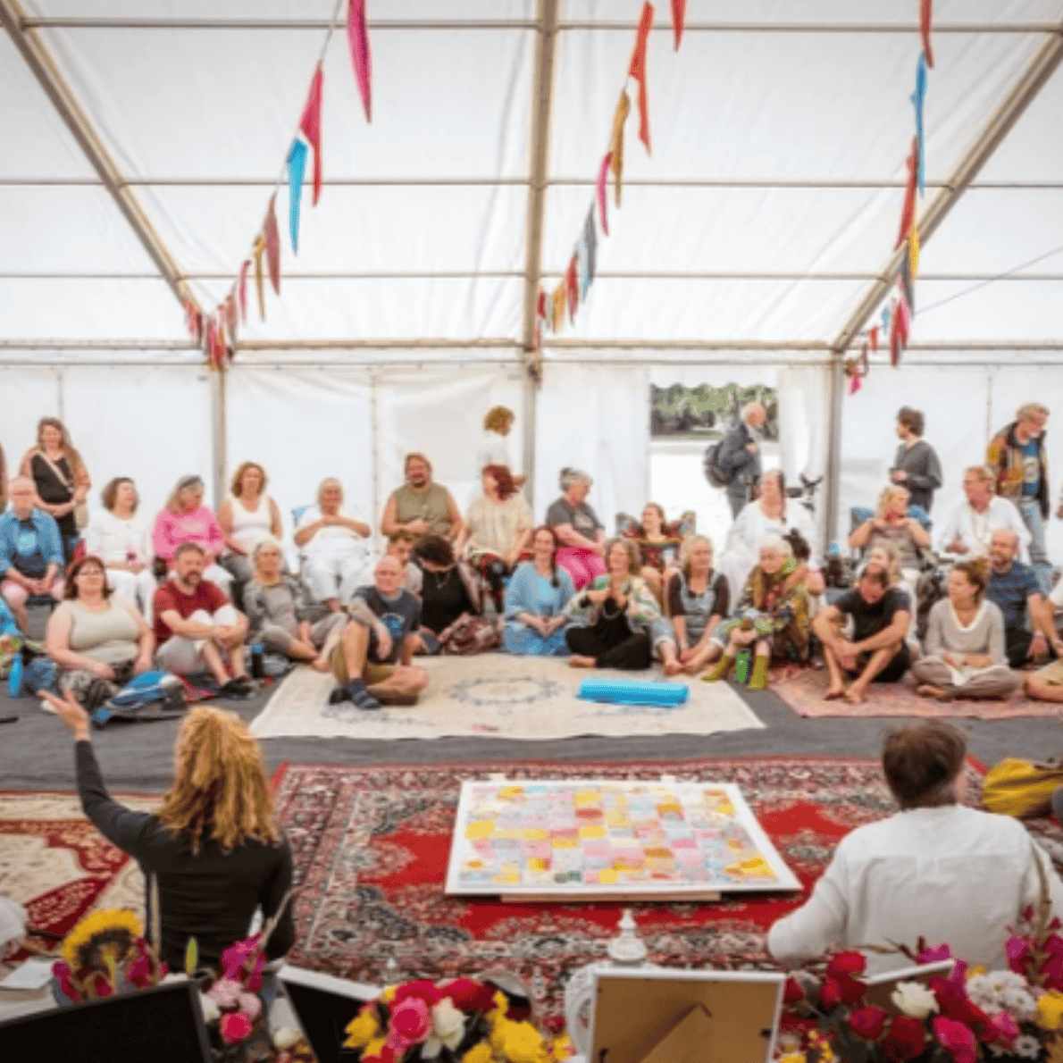 A group of people sitting on the floor inside a large tent, attending a workshop or discussion, with colorful pennant banners hanging from the ceiling.