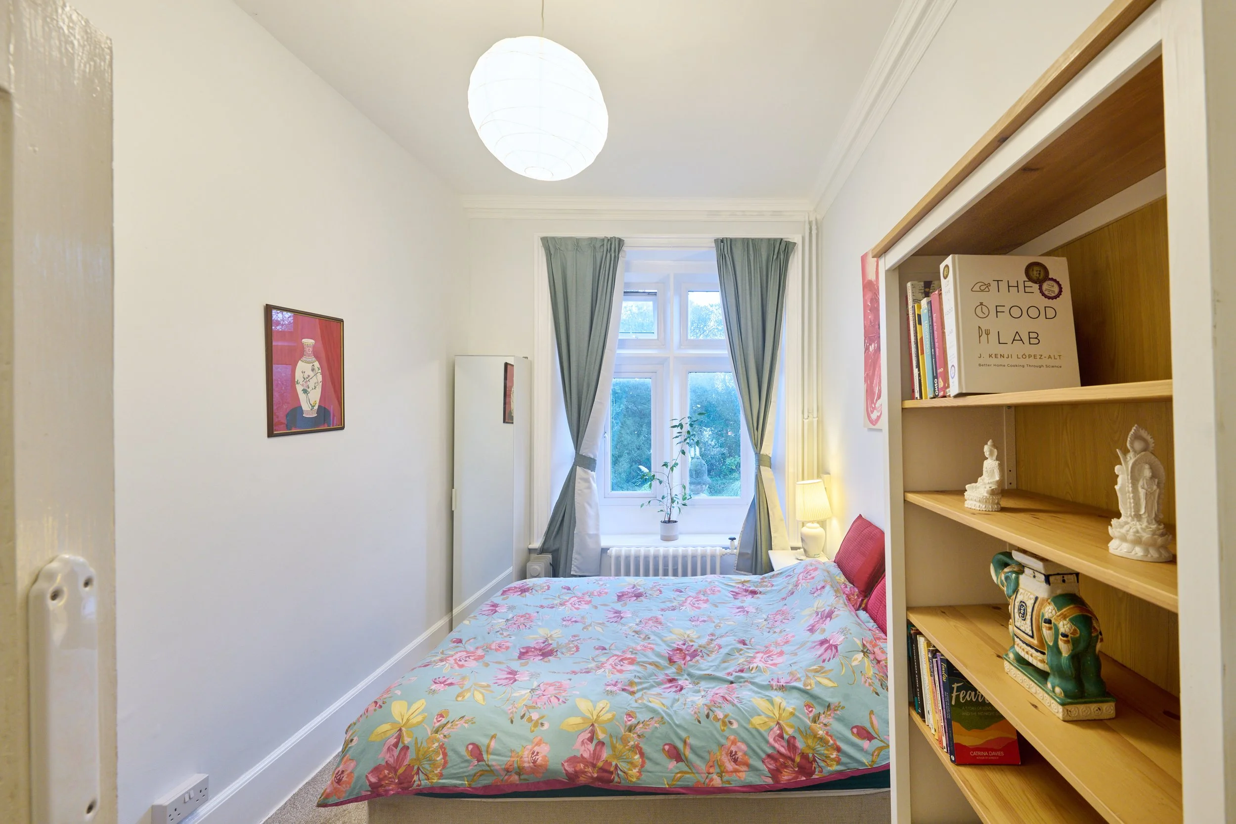 A cozy bedroom with a floral bedspread, a window with curtains, a bookshelf with various items, and framed artwork on the wall.