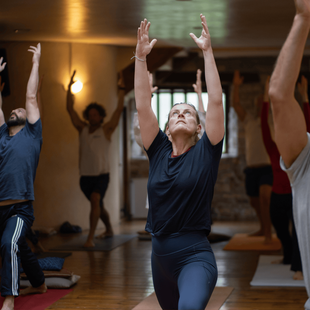 People participating in a yoga class, practicing a pose with arms raised overhead in a dimly lit indoor studio.