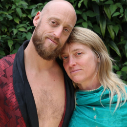 A man with a beard and a woman with blonde hair standing close together outdoors among green foliage.