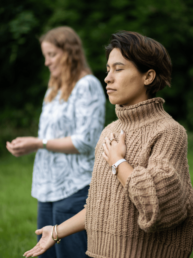 Two women practicing meditation outdoors, standing with eyes closed, hands placed on their chests, with a forested background.