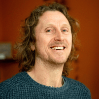 A man with curly hair, smiling, wearing a teal sweater in an indoor setting.