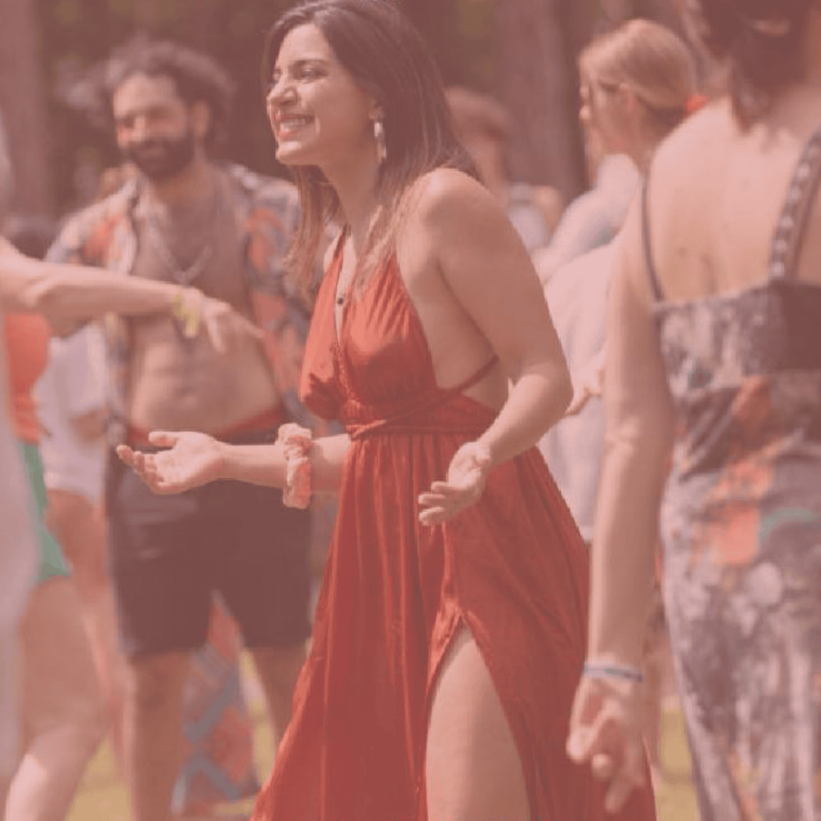 A woman in a red dress with a slit, smiling and gesturing as she socializes with other people outdoors.