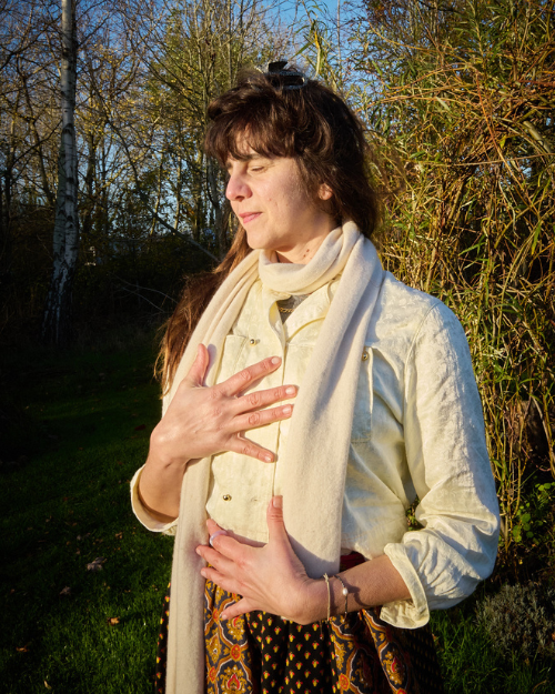 A woman practicing yoga or meditation outdoors during the daytime, standing with her eyes closed, hand on chest and stomach, in front of a natural background of trees and bushes.