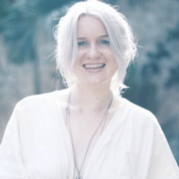 A smiling woman with short, white hair, wearing a light-colored shirt outdoors.