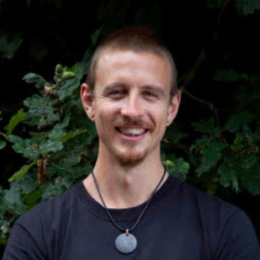 A young man with short brown hair and a beard, smiling in front of green foliage, wearing a black shirt and a necklace with a round gray pendant.