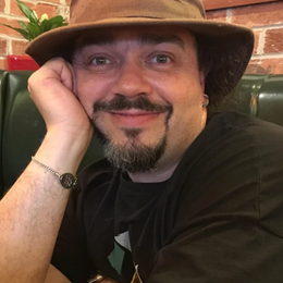 A man with a beard wearing a wide-brimmed hat, smiling, resting his chin on his hand in a restaurant or cafe setting.