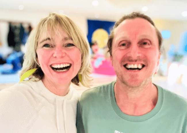 A woman with blond hair and earrings smiling next to a man with brown hair also smiling, in an indoor setting.