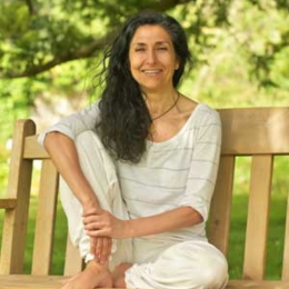 A woman with long dark hair sitting on a wooden bench outdoors, smiling, surrounded by green trees and grass.