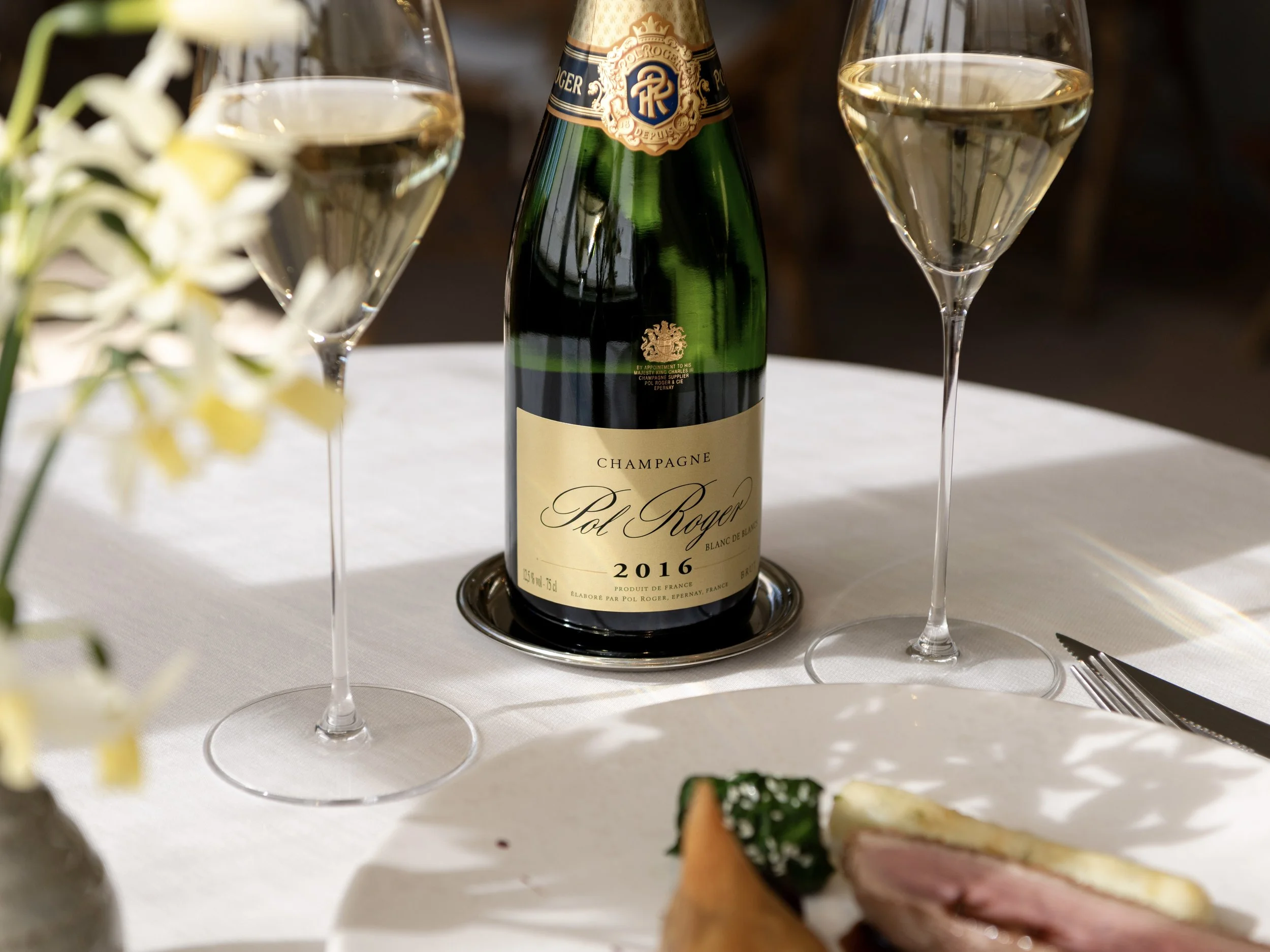 A bottle of Pol Roger 2016 Vintage Champagne on a white tablecloth, flanked by two filled flutes and a plate of gourmet food. Strong natural sunlight illuminates the table at The Woodcote restaurant.