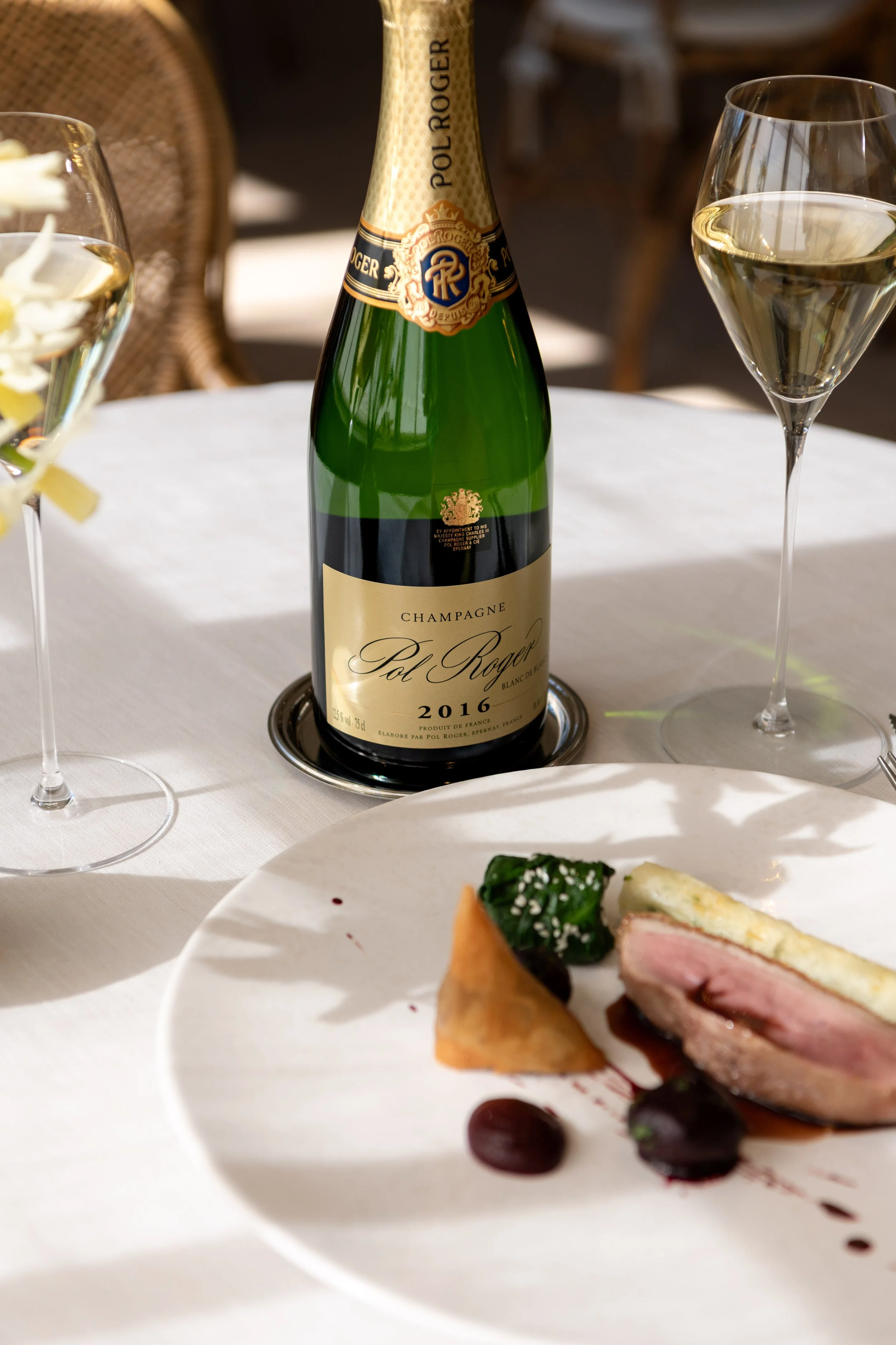 A bottle of Pol Roger 2016 Champagne with two glasses and a gourmet duck dish on a sunlit table at The Woodcote restaurant.