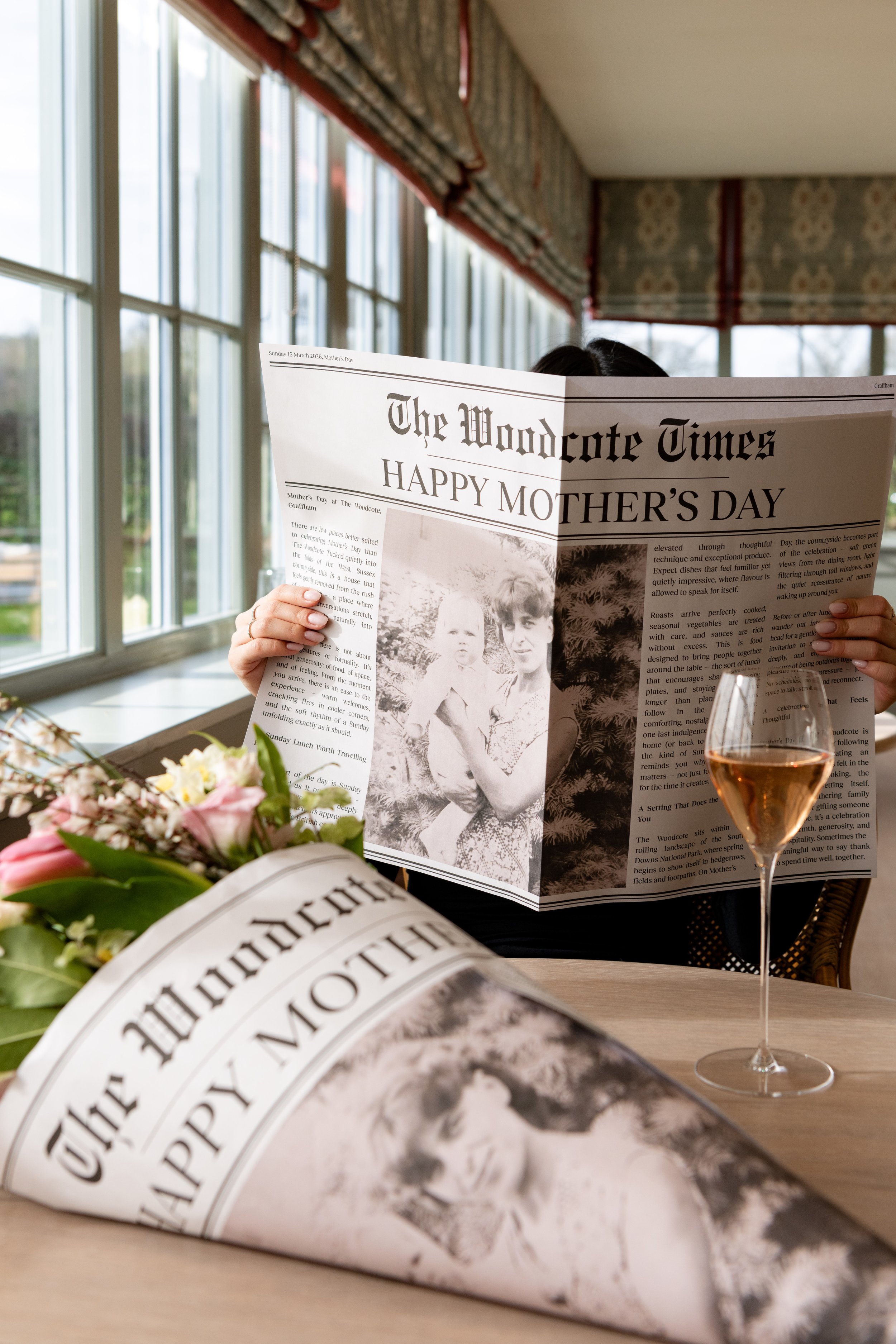 A person reading 'The Woodcote Times' Happy Mother's Day special edition newspaper at a restaurant table with a flower bouquet and a glass of wine