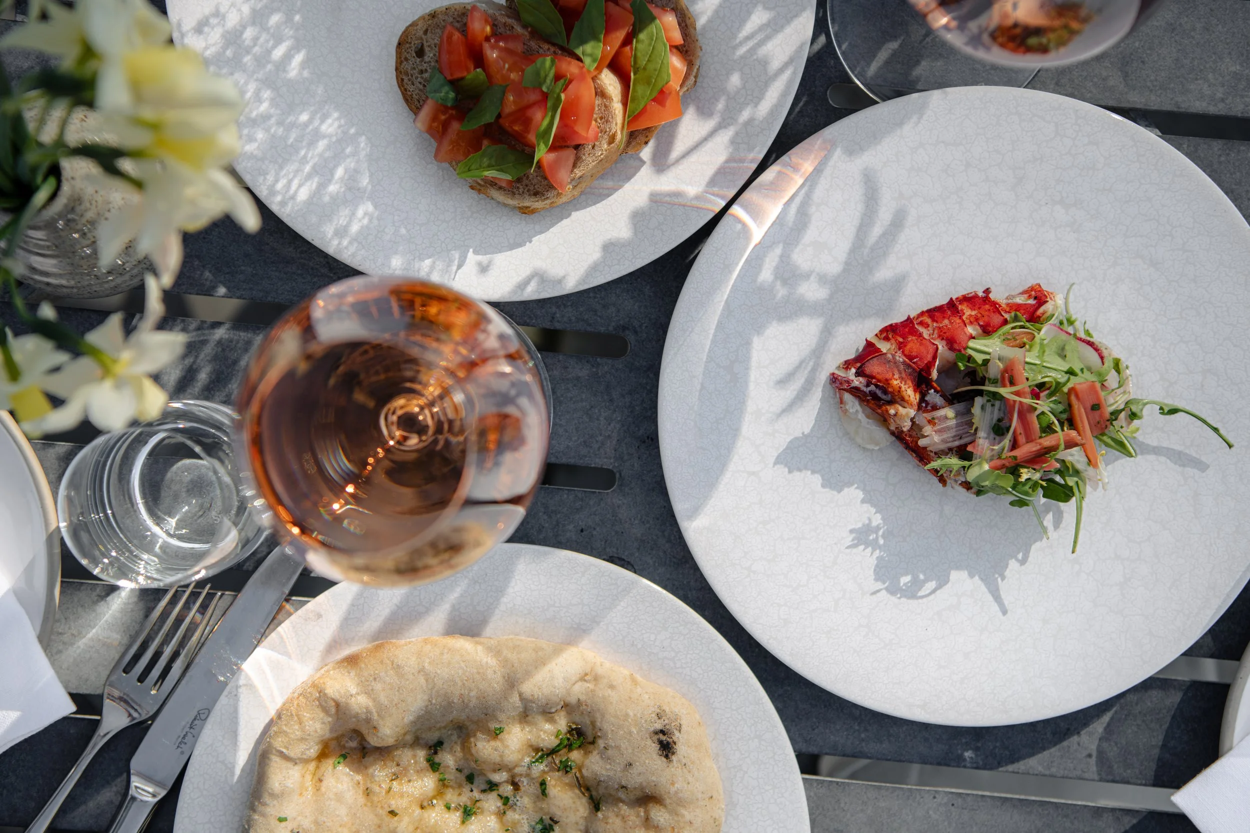 An aerial view of gourmet starters at The Woodcote terrace, featuring lobster salad, tomato and basil bruschetta, and flatbread paired with glasses of chilled rosé wine.