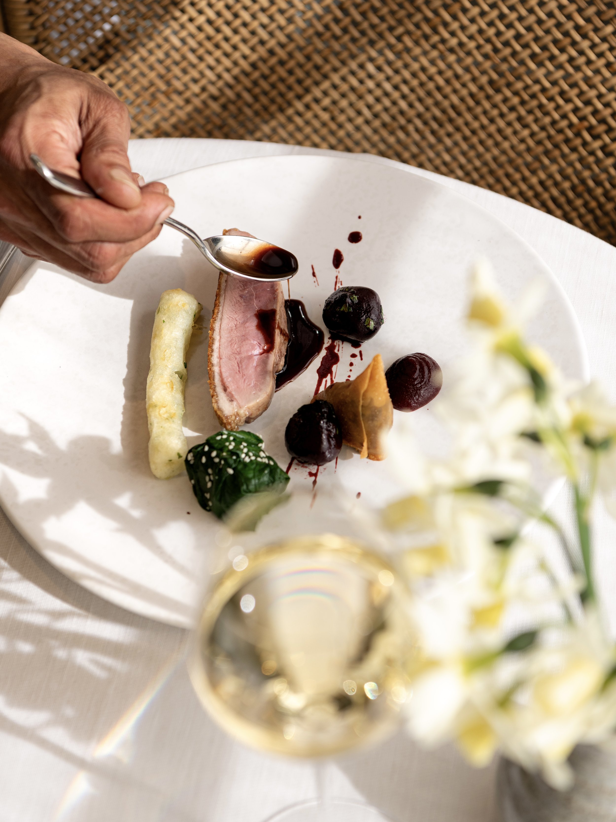 A hand pouring rich jus over a perfectly cooked duck breast. A blurred glass of Pol Roger champagne sits in the foreground on a sunlit table at The Woodcote.