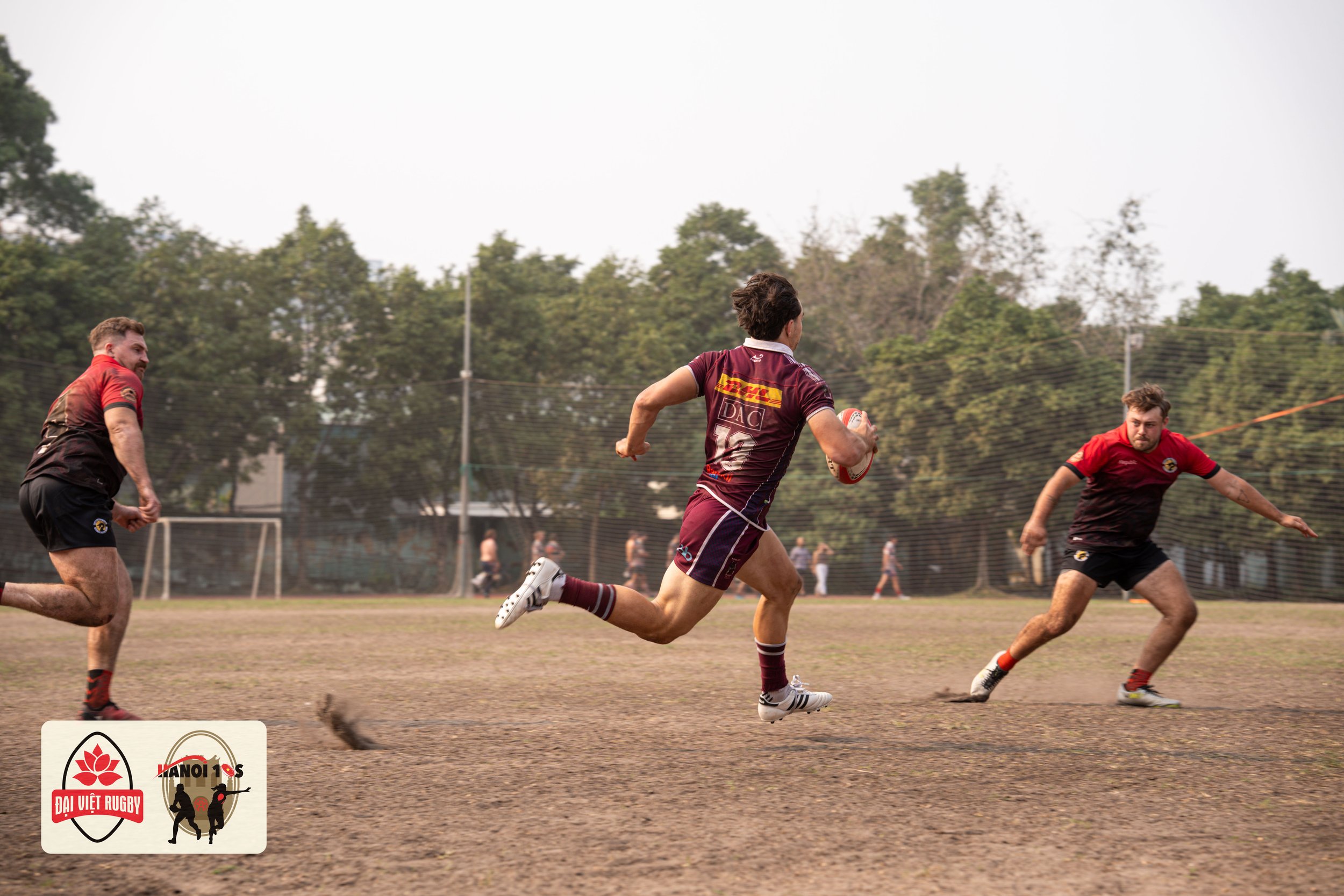 Rugby players running on a dirt field, with one holding a rugby ball, during a game. Two players in red and black uniforms and another player in maroon and white uniforms. Goals and trees in background.