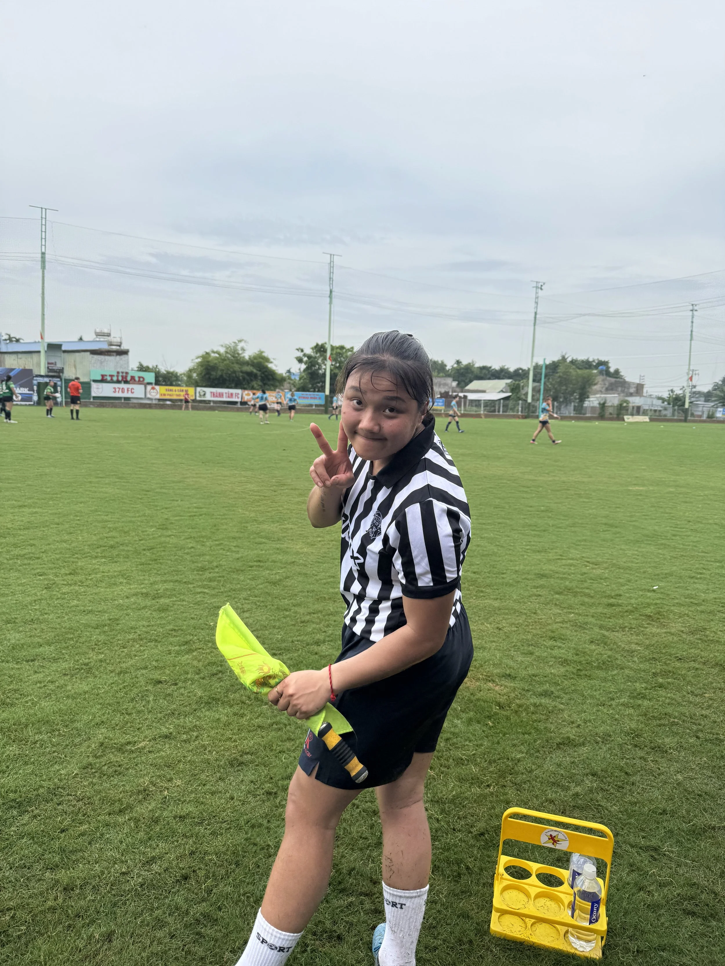 A young female rugby match offical, making a peace sign with her right hand while holding a yellow sports cone in her left hand. She is smiling and wearing a black and white striped jersey and black shorts, with white socks and sneakers.