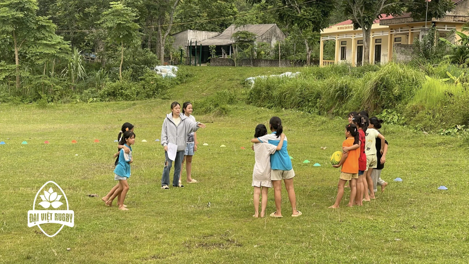 Children and two female coaches standing on a grassy field, engaged in a rugby & life skills practice session. One coach is instructing the kids, who are hugging a rugby ball. The scene is outdoors on a cloudy day, with trees in the background.
