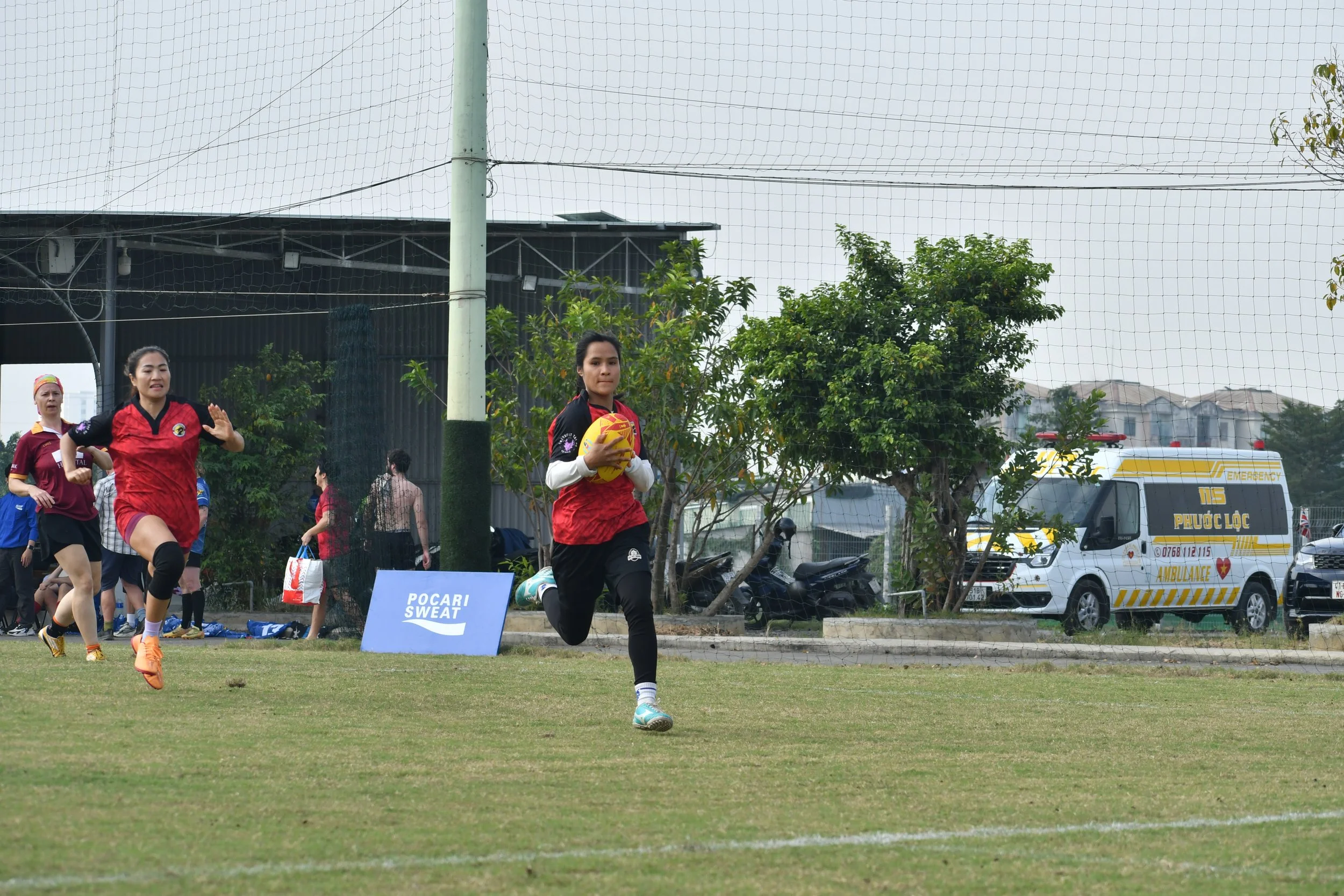 A female player from the Hoa Binh Rugby Club is holding a yellow ball runs on a grassy field during a game, on her way to scoring one of many tries in the finals matches. Other players and an emergency ambulance vehicle in the background.