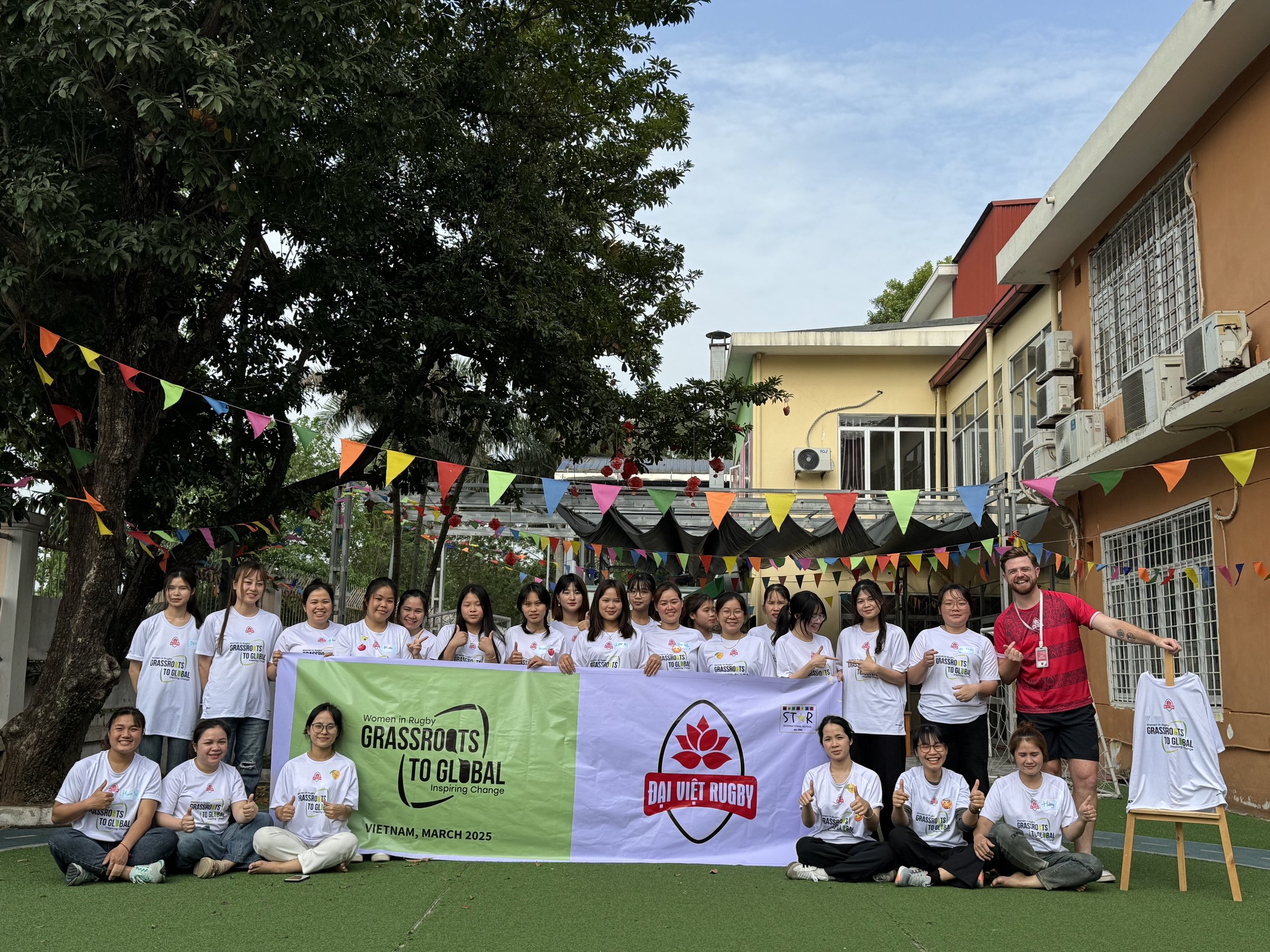 Group of women and a man holding banners and posing outdoors with colorful flags, trees, and buildings in the background.