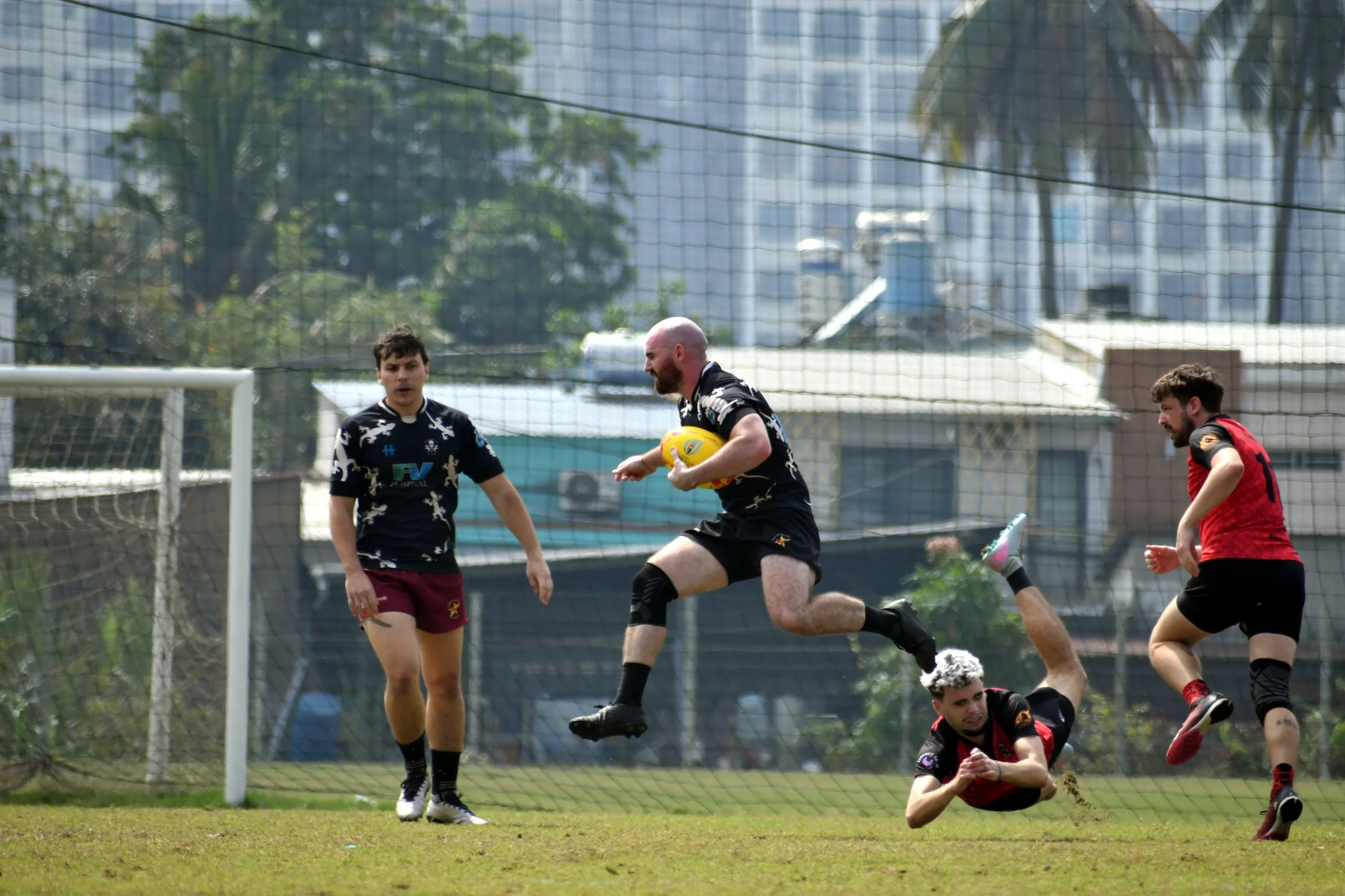 Rugby players in black and red jerseys playing on a grassy field, with one player jumping in the air holding a yellow rugby ball, while another player is on the ground and others are nearby.