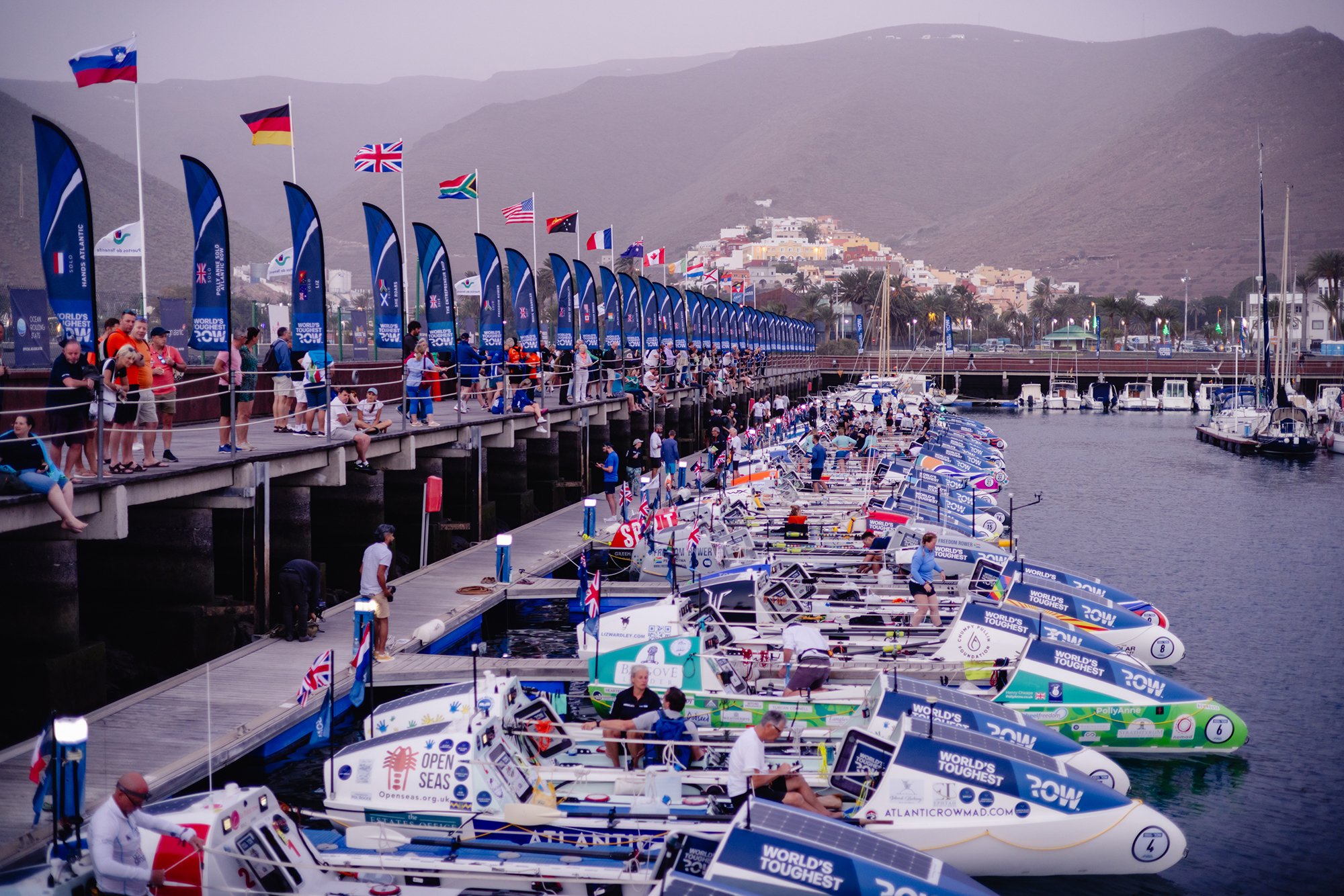 La Gomera marina with World's Toughest Row boats lined up, each featuring sponsor logos and text such as "World's Toughest Row." Flags fly above the pier, which is crowded with people watching the event.