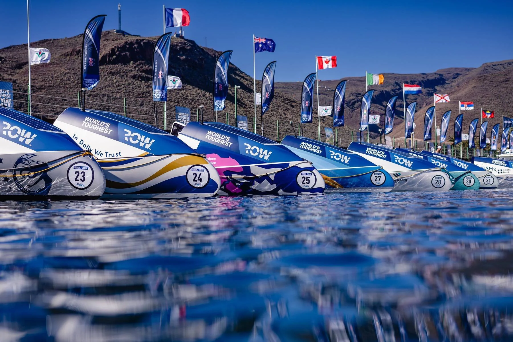 Rowing boats lined up in water, displaying "World's Toughest Row" branding, with various international flags in the background.