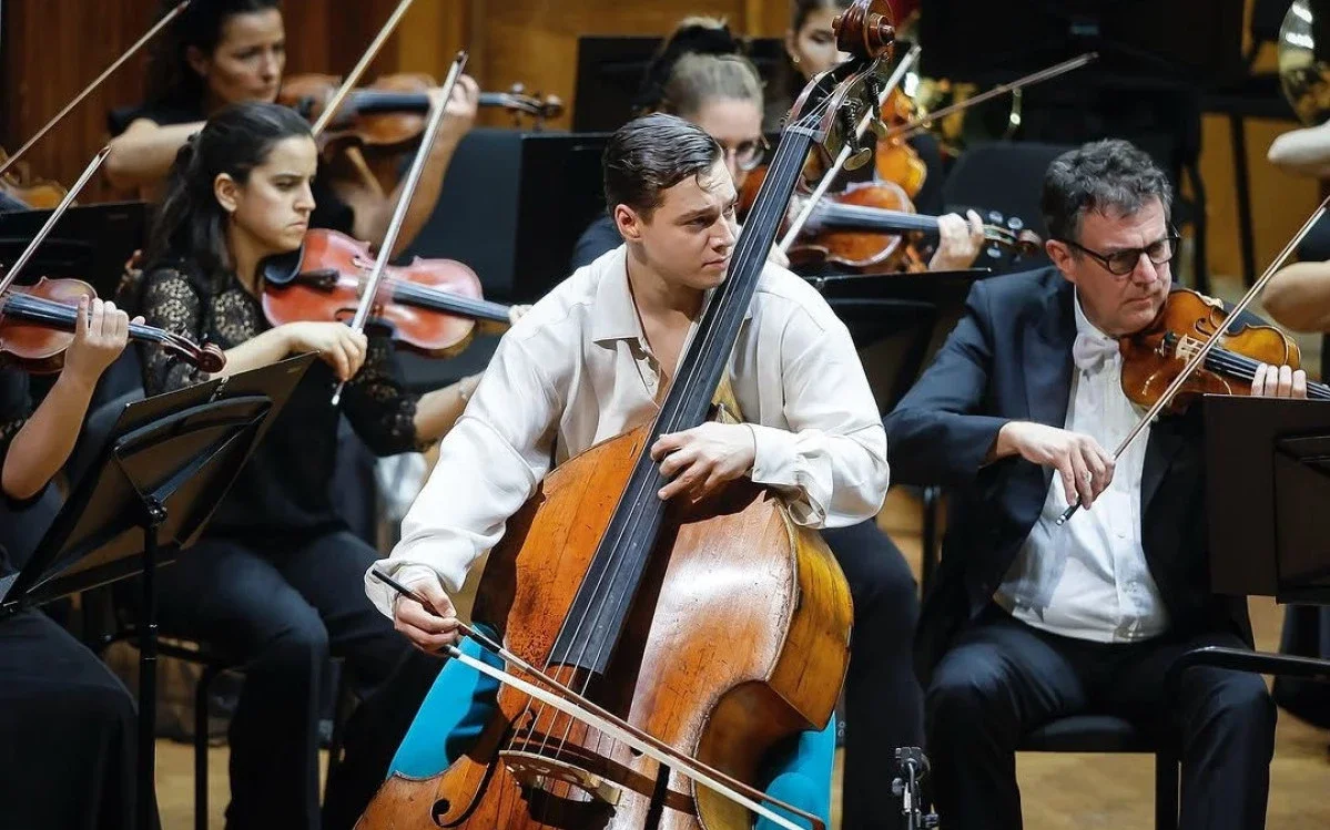 Orchestra musicians playing string instruments during a performance, with a focus on a young man playing a double bass and an older man playing a violin.