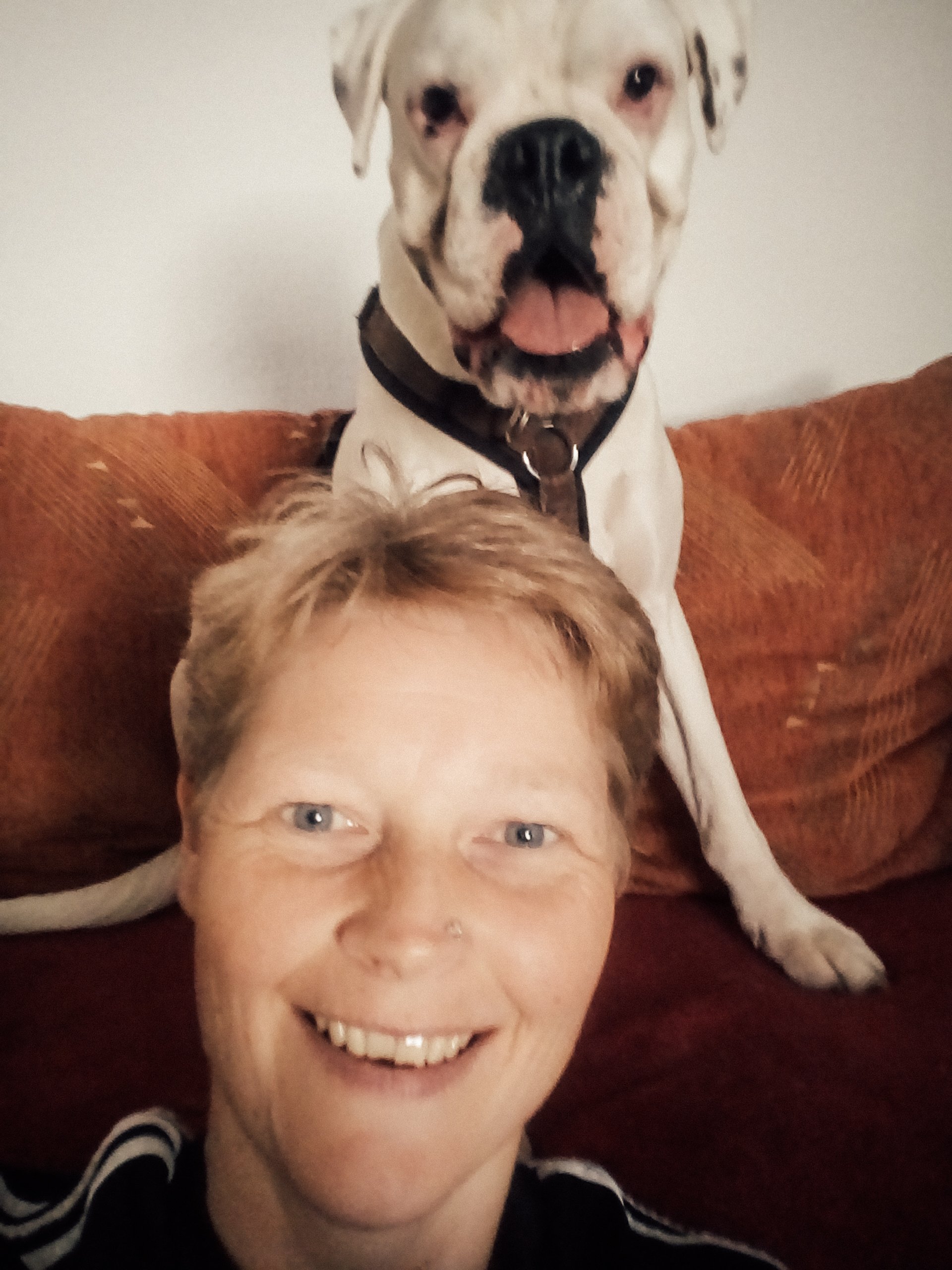 Eine lächelnde Person macht ein Selfie, während im Hintergrund ein weißer Boxerhund auf einem Sofa sitzt.