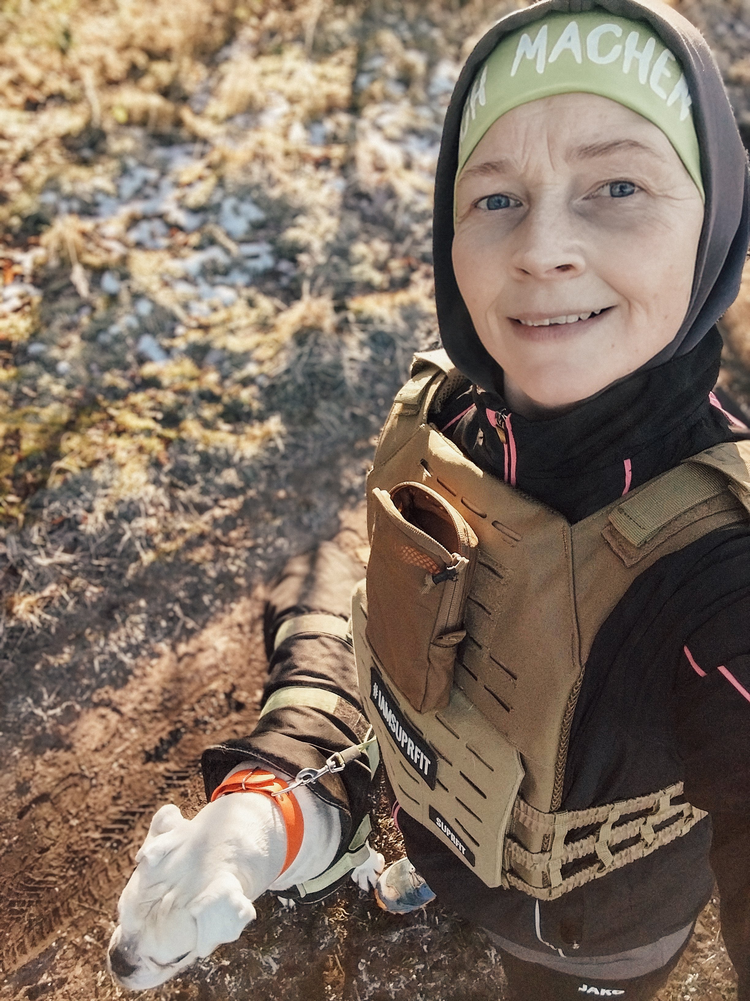 Person in Sportkleidung und Mütze macht ein Selfie im Freien, während ein weißer Hund an der Leine neben ihr steht.
