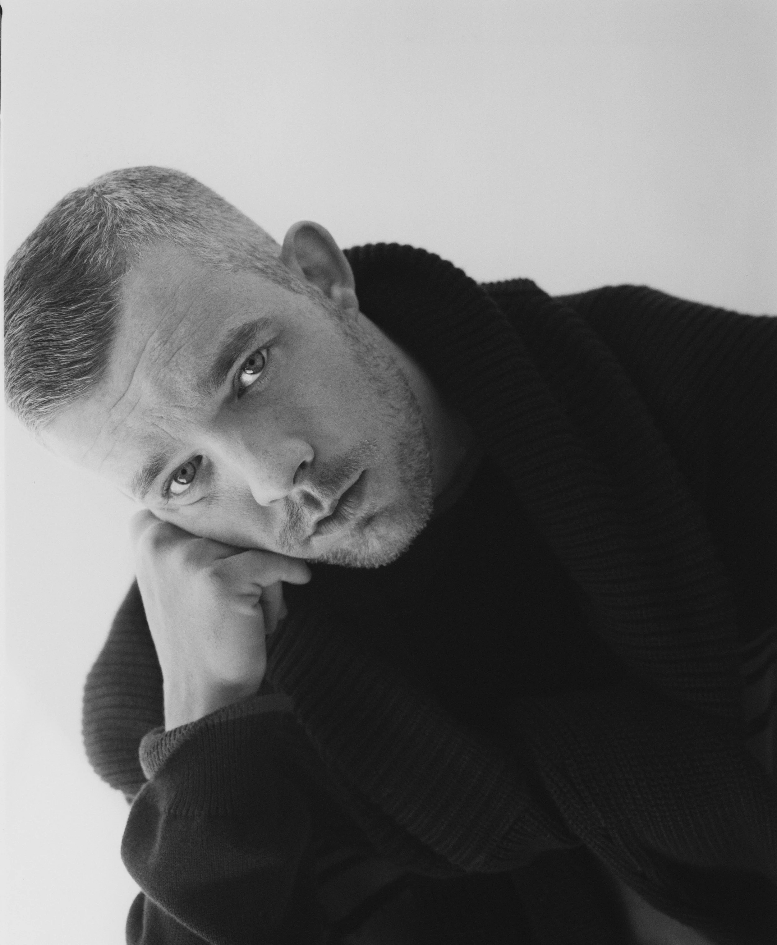 A close-up black and white portrait of a man with short hair and a beard, resting his face on his hand, wearing a dark sweater.