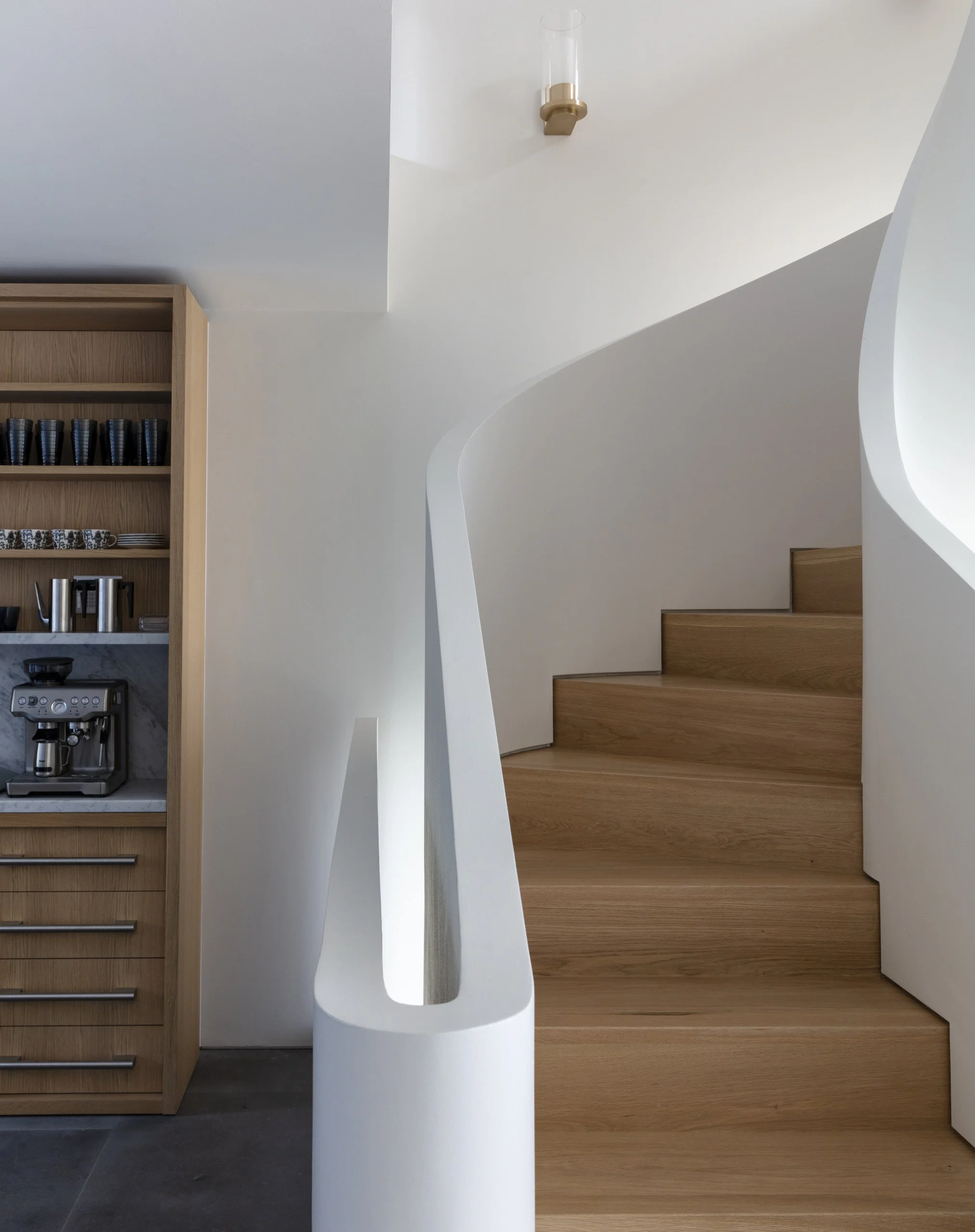 Interior view of modern staircase with wooden steps, white curved walls, and a black and grey coffee maker on a wooden shelf.