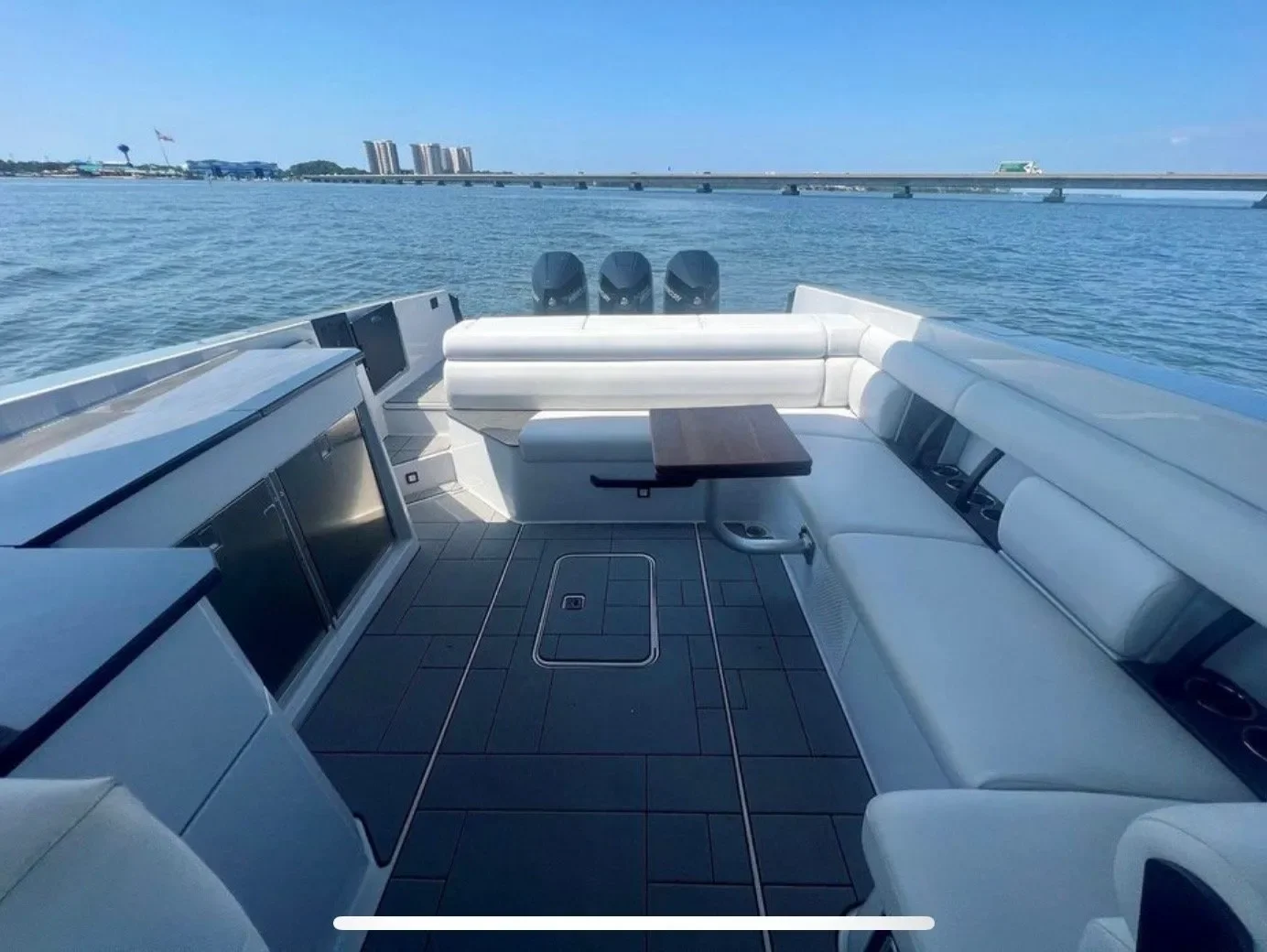 View of a yacht deck with white cushioned seating and a small wooden table, overlooking the water and city skyline and bridge in the distance.