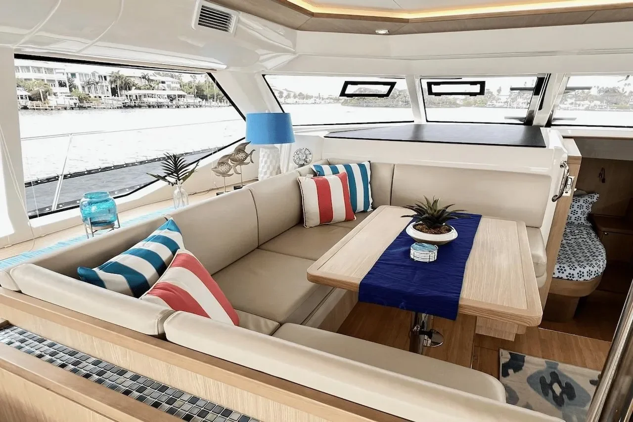 Interior of a yacht with a beige L-shaped sofa, colorful striped pillows, a wooden table with a blue runner and a potted plant, decorative fish sculptures, and windows with a view of water and houses.