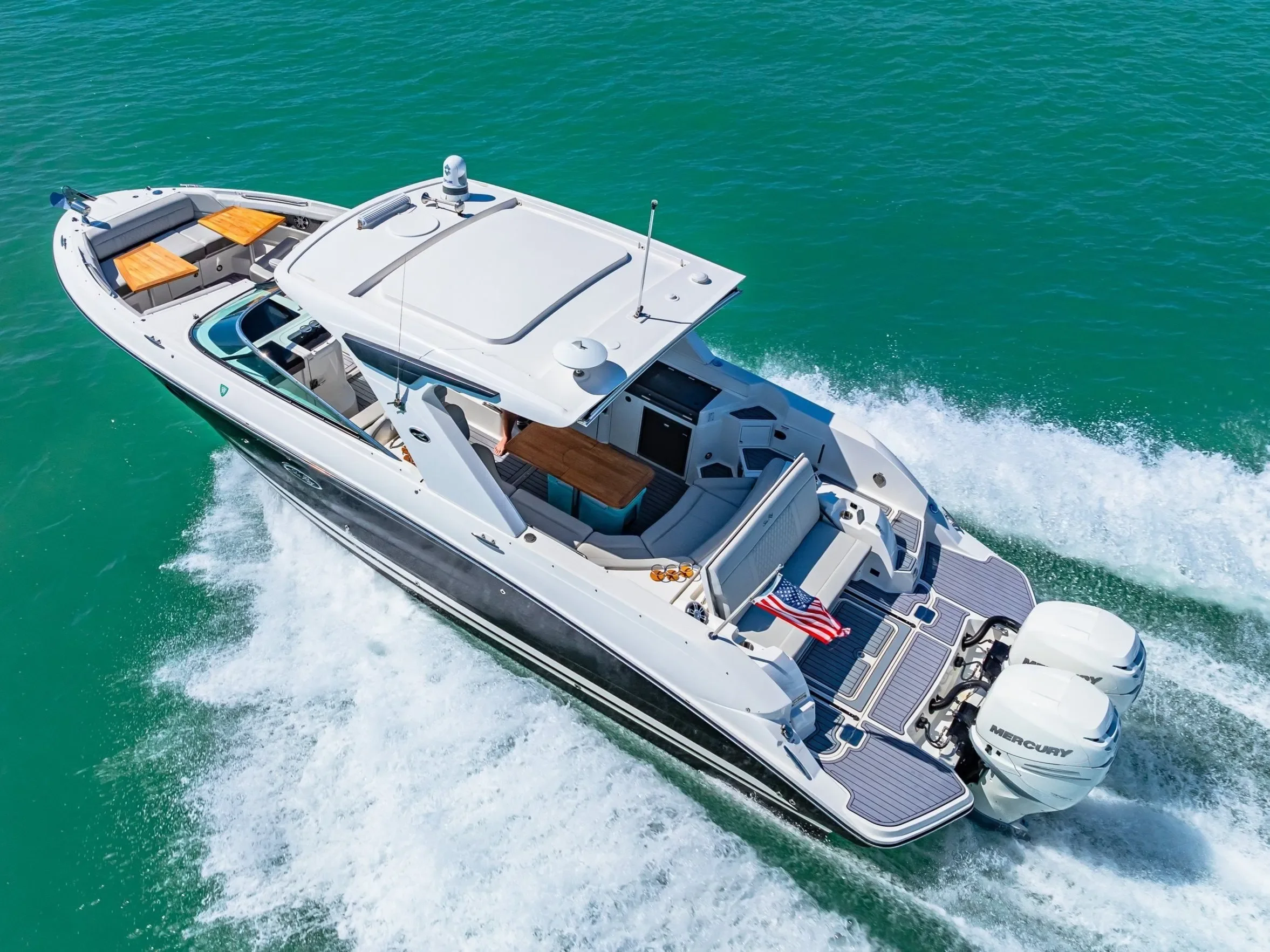 kokomo charters - 38' Top Shelf - An aerial view of a white luxury motor yacht cruising on turquoise water, with seating areas, a wooden table, an American flag, and twin Mercury engines at the stern.