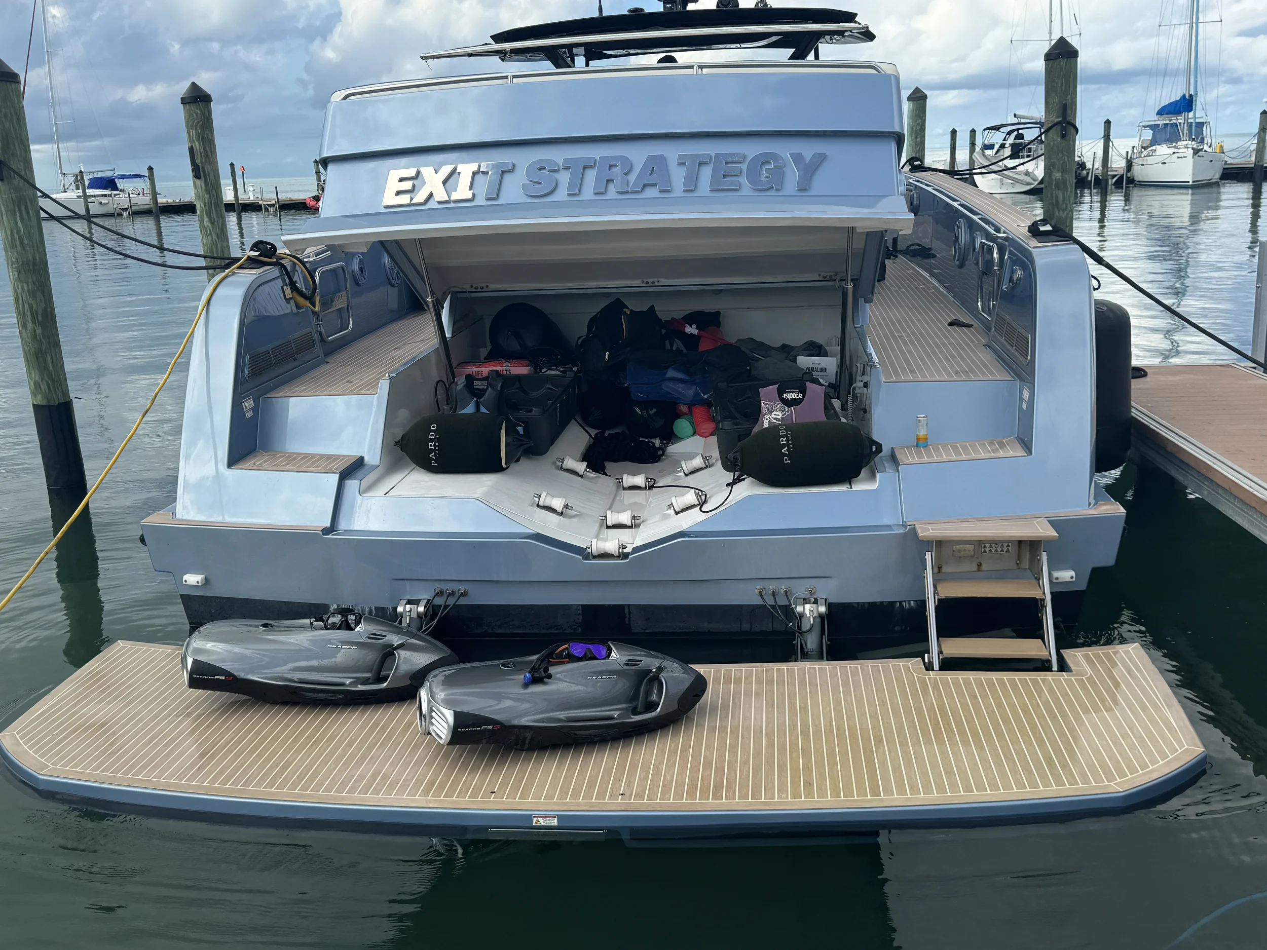 A modern yacht with the name 'EXIT STRATEGY' docked at a marina, with watercraft inside and two jet skis on the deck at the stern.