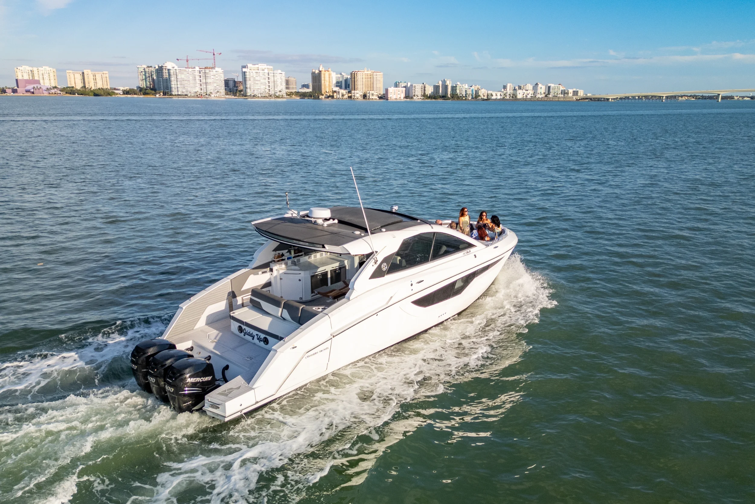Kokomo charters - bachelorette boat party Sarasota - A white yacht sailing on the water with people relaxing on the deck, and a city skyline with tall buildings in the background.