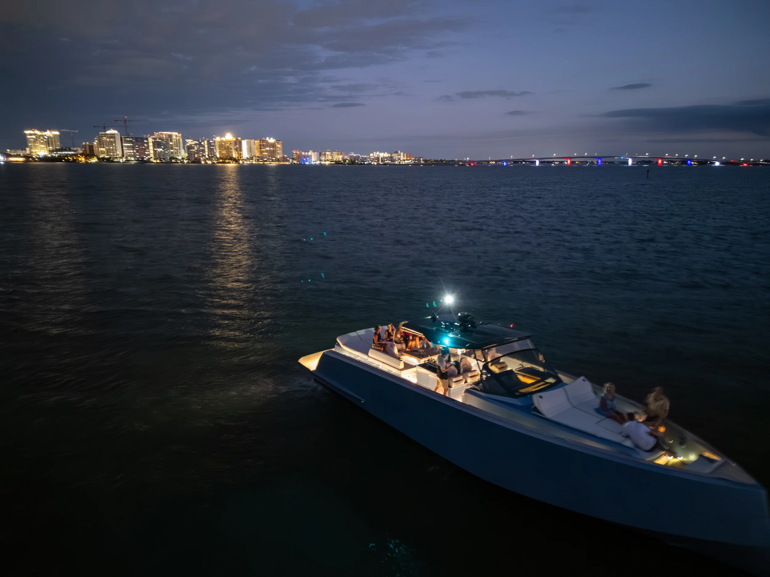 Kokomo charters - yacht float plan Sarasota - A boat cruising on water during nighttime with city lights and a bridge in the background.