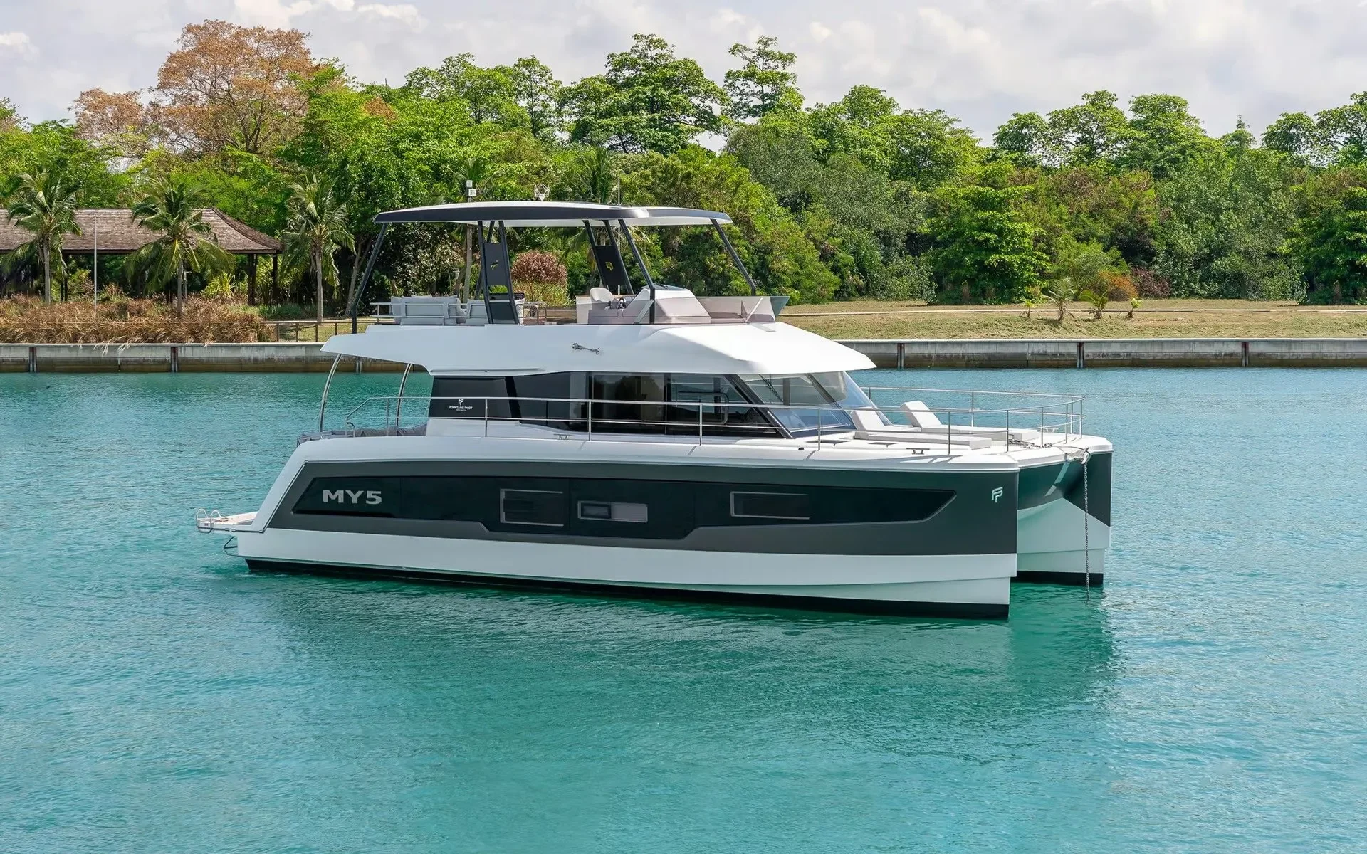 A modern white and black yacht named MY5 floating in calm turquoise water near a tropical shoreline with lush green trees and a small building in the background.