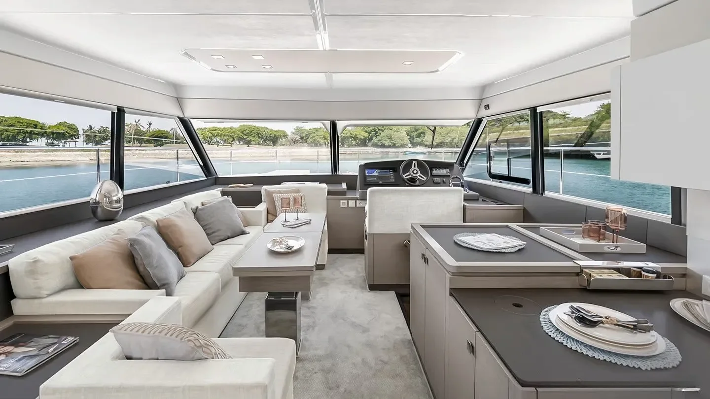 Interior of a yacht's living area with large windows showing water and trees outside, featuring a white sofa with beige and gray pillows, a beige armchair, a sunken console, and a dashboard with steering wheel and instruments.