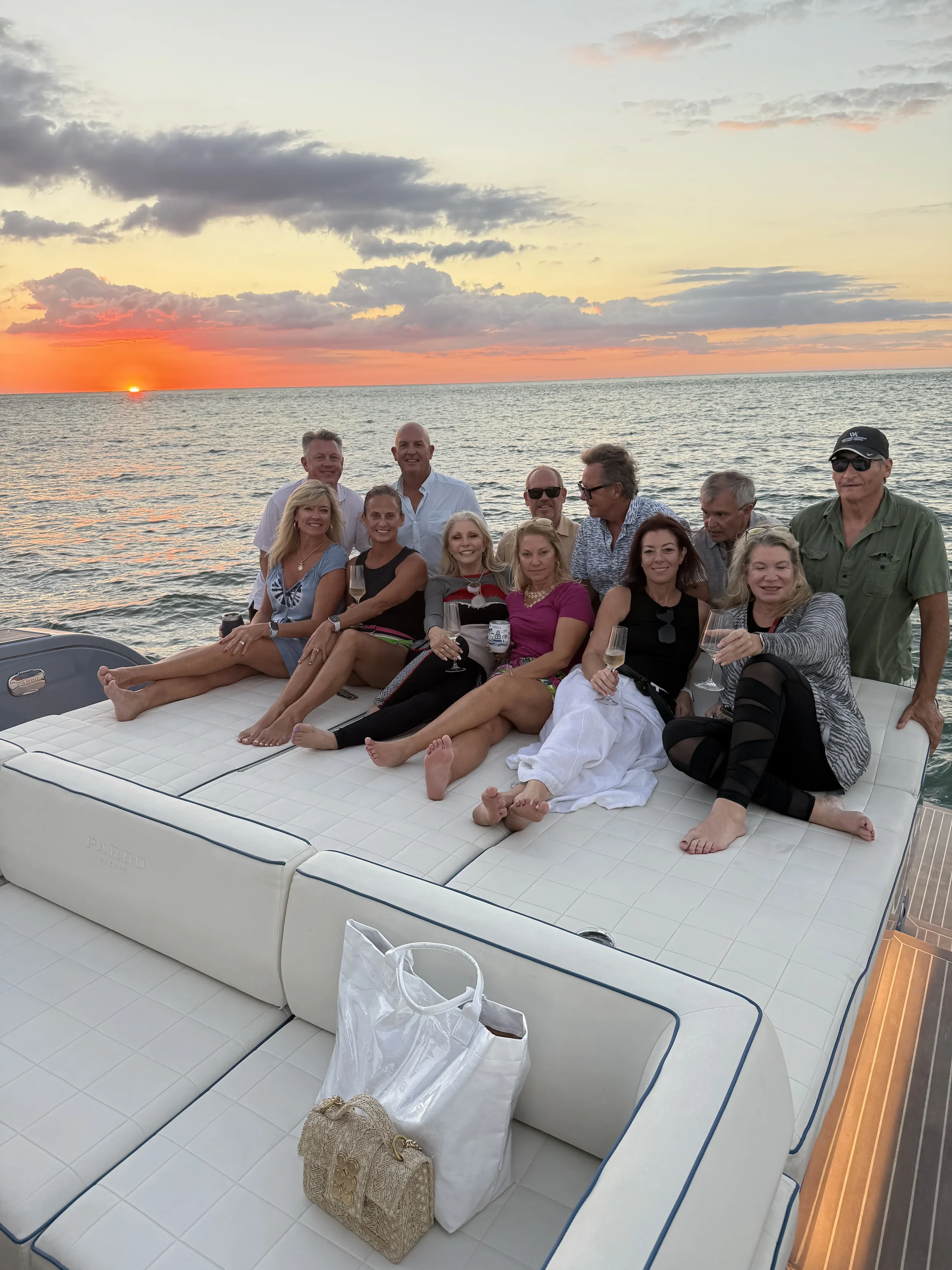 A group of twelve people sitting on the deck of a yacht during sunset, holding drinks and smiling.
