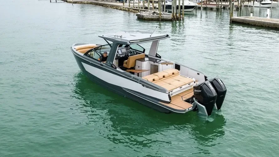 A modern motorboat with a white and black hull, tan leather seating, and twin outboard engines is floating in a marina with docks and boats in the background.