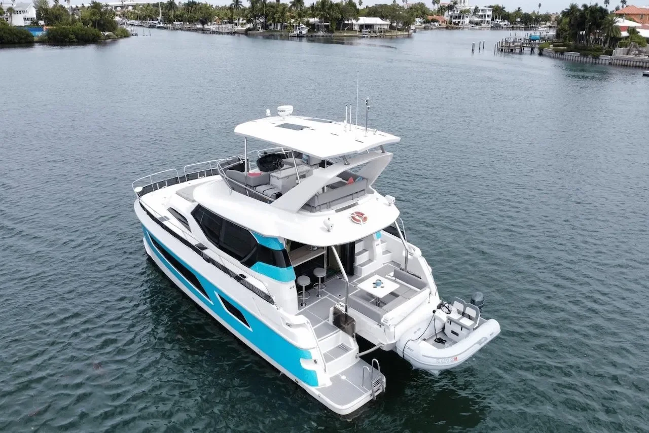 A modern white yacht with blue accents floating on calm water near a shoreline with residential houses and palm trees.