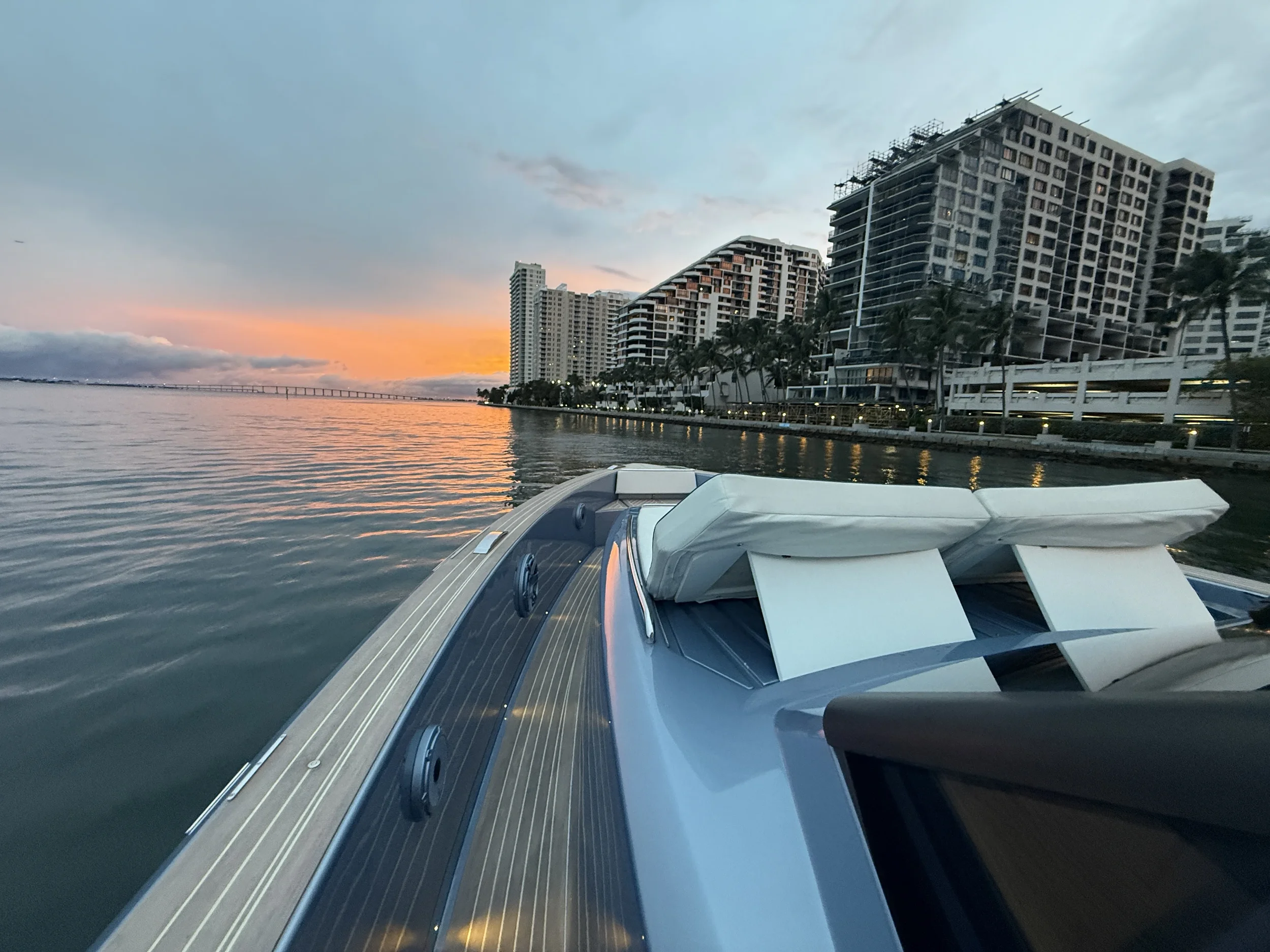 View from a boat on calm water during sunset, showing high-rise buildings along the shoreline and a bridge in the distance, with a partly cloudy sky.
