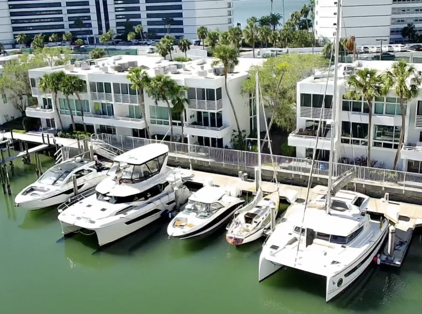 Luxury yachts and boats docked at a marina in front of modern waterfront apartments with palm trees.