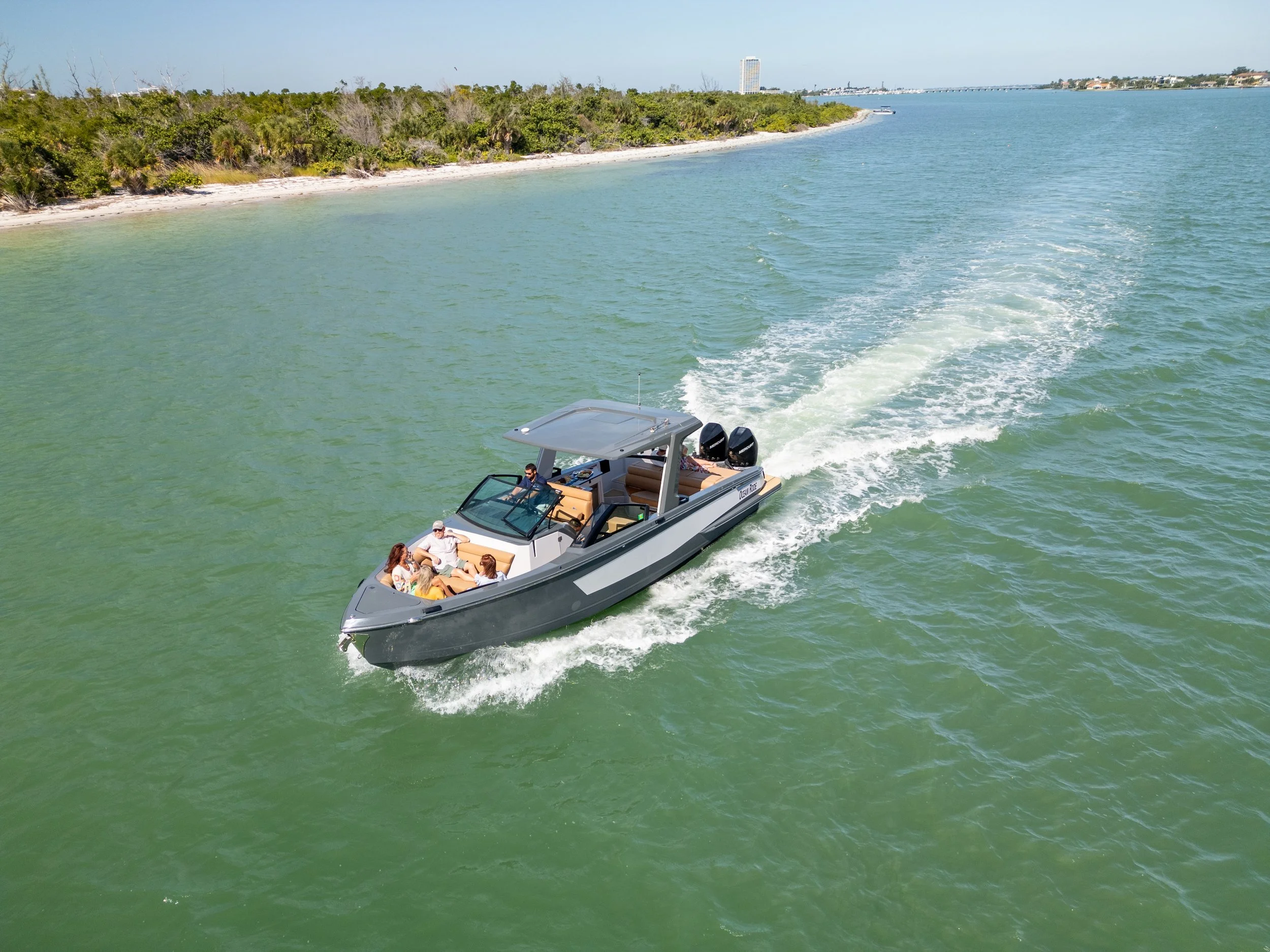 Kokomo charters - bachelorette boat party Sarasota - A group of five people on a sleek gray motorboat cruising through green water near a coastal area with trees and buildings in the background.