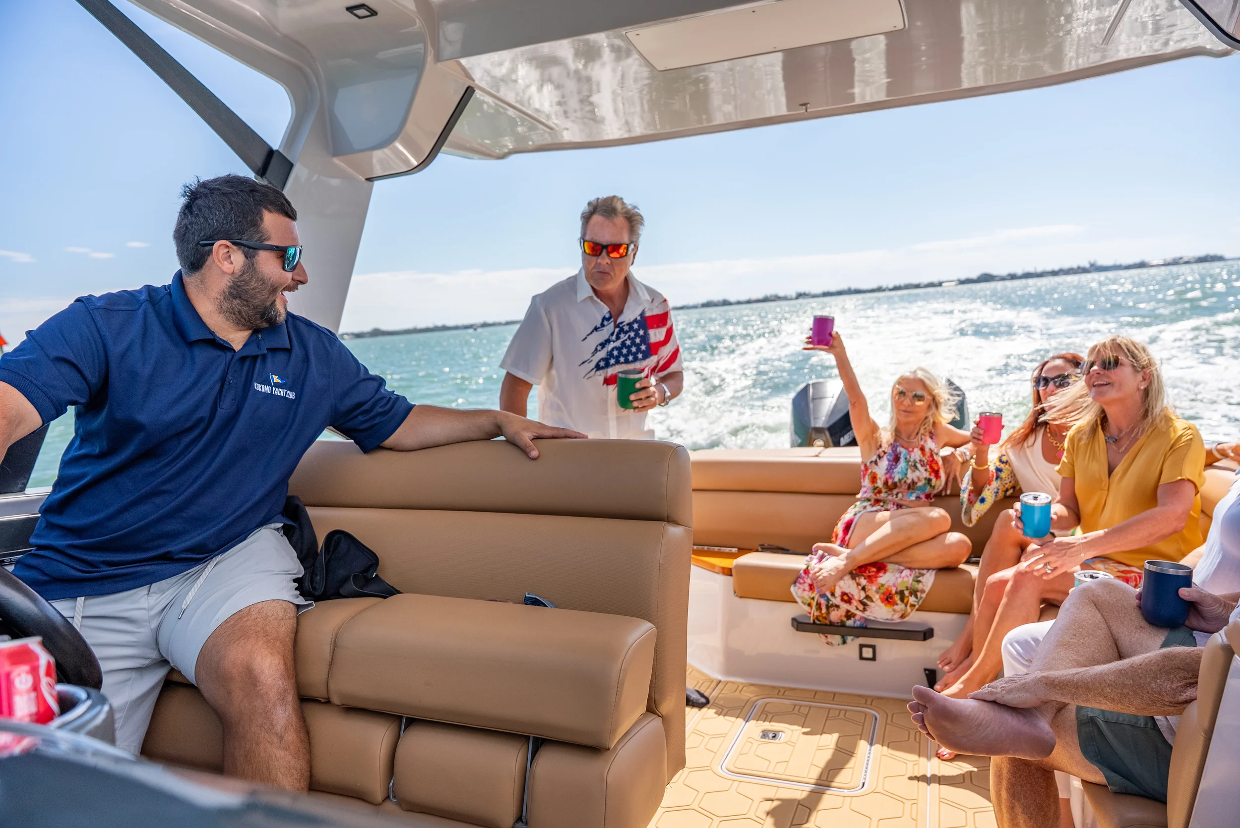 Kokomo charters - Lido Key yacht tours - Group of friends enjoying drinks on a boat, with a man in a blue polo shirt sitting and three women standing and sitting, all holding colorful cups, on a sunny day with water in the background.