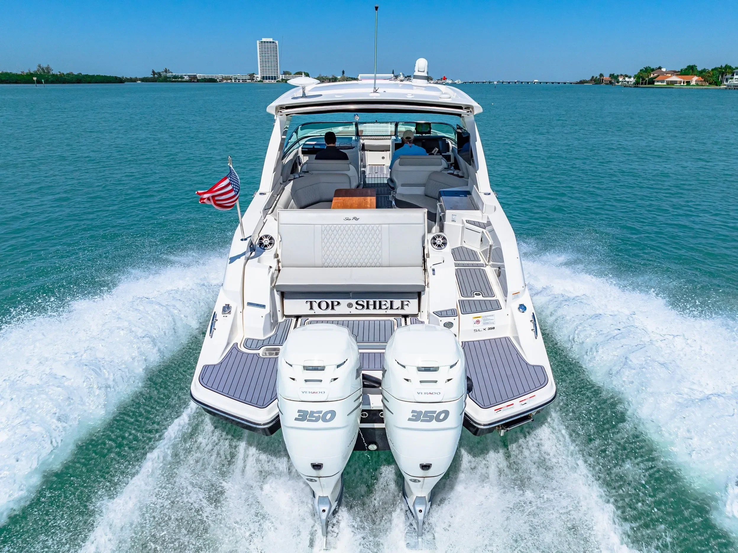 A white motor yacht with twin outboard engines speeds across a turquoise body of water, creating a wake, with a cityscape and bridge in the background.